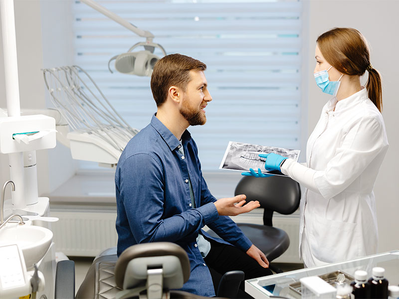 Junger Mann sitzt im Zahnarztstuhl, Zahnärztin zeigt ihm ein Röntgenbild in moderner heller Praxis.