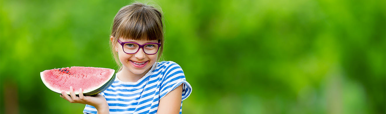 Mädchen mit Brille und Zahnspange isst im Garten fröhlich eine große Scheibe Wassermelone.