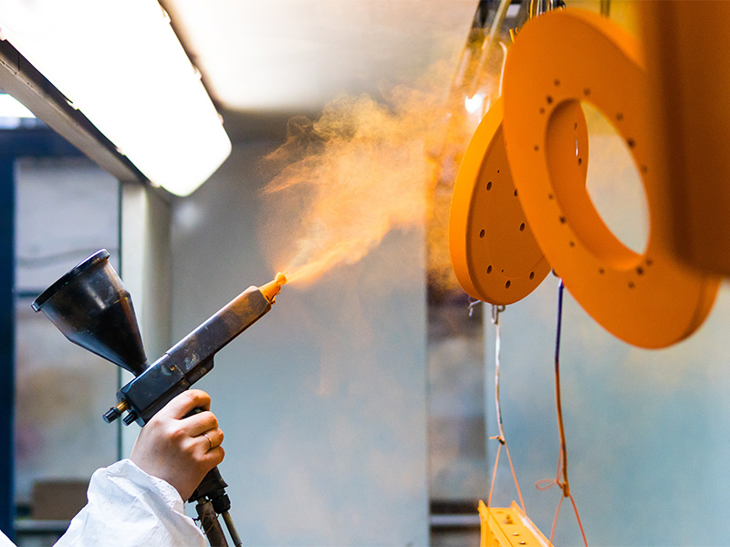 Mitarbeiter in Schutzkleidung beschichtet Metallteile mit einer Sprühpistole und trägt Pulverlack gleichmäßig auf.